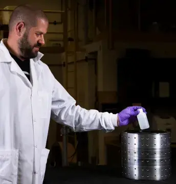 A young man on the left is dressed in white lab coat and wears purple gloves; he has very low haircut and has a beard and mustache. He is in profile facing the right. He has outstretched his left arm and holds a small white plastic. In the dark, blurry background could be seen lab furniture