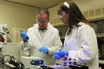 Two scientists, a female with long hair on the right and a male starting to go bald, dressed in white lab coats and blue gloves and protective goggles work in a lab. The woman is watching the man’s hands as he is holding a pipette and a small test tube. Infront of them there is lab machinery. The room is off white and in the background there are vents and glass.