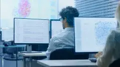 A man looks at two computer screens; a woman looks at a computer screen behind him. 